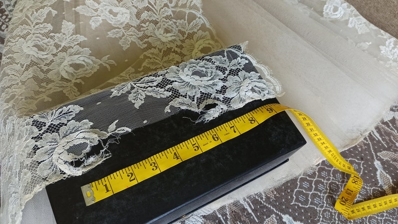 Op de afbeelding: Ivoor kanten stof met bloempatronen, een zwarte doos en een gele meetlint. De tape geeft een meting van ongeveer 23 cm aan. Het kant is over de doos gedrapeerd.