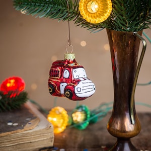 May include: A red and white fire truck Christmas ornament with a glossy finish, hanging from a wire hook. The ornament features black wheels, a white front grille, and a red body. It is suspended near a copper-colored vase and pine branches.