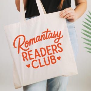 May include: Cream-colored tote bag with the words "Romantasy READERS CLUB" in orange script. The bag has two handles and is held up by a person. The text includes two small heart symbols. The bag is made of a sturdy fabric.