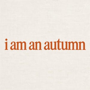 May include: A square graphic print featuring the text "i am an autumn" in a bold, burnt orange serif font. The lettering is centred against a light, off-white textured background that resembles the weave of a piece of fabric or canvas.