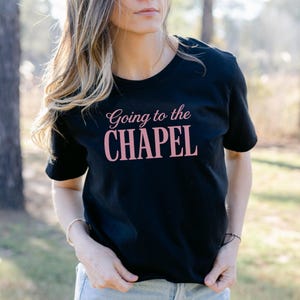 Puede incluir: Una camiseta negra de manga corta con el texto Going to the CHAPEL impreso en color rosa claro en el pecho. La prenda cuenta con un cuello redondo y un corte est&aacute;ndar, ofreciendo un dise&ntilde;o sencillo y claro para uso cotidiano.