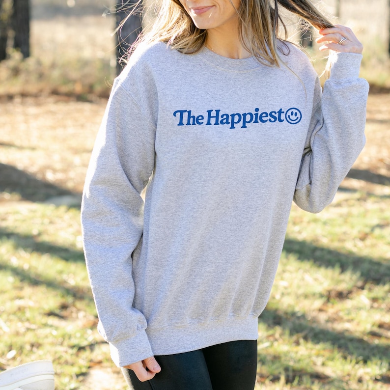 Puede incluir: Una sudadera gris claro de cuello redondo con el texto The Happiest en una fuente serif azul oscuro, acompa&ntilde;ado de un peque&ntilde;o icono de cara sonriente azul al final del texto. La prenda tiene mangas largas y pu&ntilde;os y dobladillo acanalados.