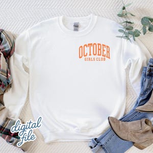 May include: A white long-sleeve crewneck sweatshirt featuring the orange text OCTOBER GIRLS CLUB printed in a collegiate font on the upper left chest area. The garment has a ribbed collar, cuffs, and hem, and is displayed on a textured surface.