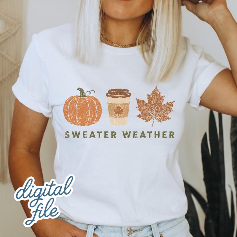 Puede incluir: Una camiseta blanca de manga corta con un estampado gr&aacute;fico de una calabaza naranja, un vaso de caf&eacute; beige y una hoja de arce marr&oacute;n. Debajo de las ilustraciones, el texto "SWEATER WEATHER" est&aacute; impreso en un estilo de fuente verde oscuro.