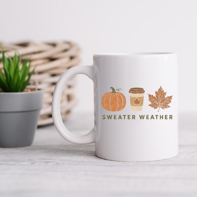 Puede incluir: Una taza de cer&aacute;mica blanca con un estampado gr&aacute;fico de una calabaza naranja, una taza de caf&eacute; color canela y una hoja de arce marr&oacute;n. Debajo de las ilustraciones, el texto SWEATER WEATHER est&aacute; impreso en letras verdes.