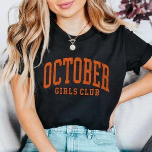 May include: A black short-sleeved t-shirt featuring the text OCTOBER GIRLS CLUB in a distressed orange font across the chest. The shirt is paired with light blue denim jeans and a gold-toned necklace with a circular pendant.