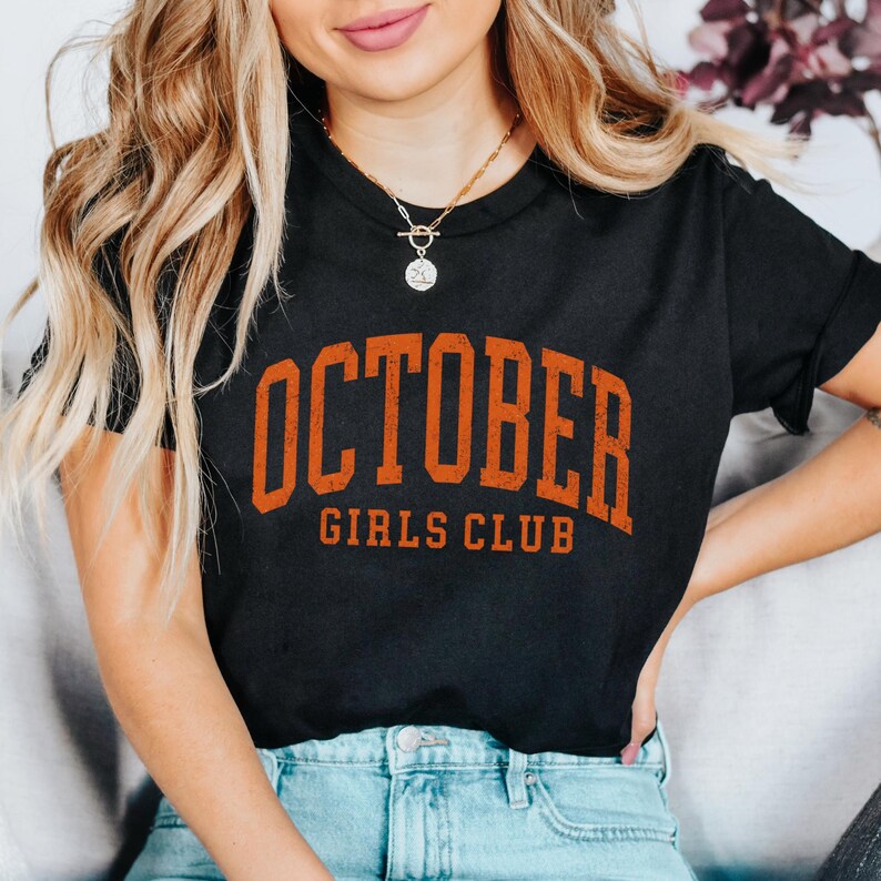 May include: A black short-sleeved t-shirt featuring the text OCTOBER GIRLS CLUB in a distressed orange font across the chest. The shirt is paired with light blue denim jeans and a gold-toned necklace with a circular pendant.