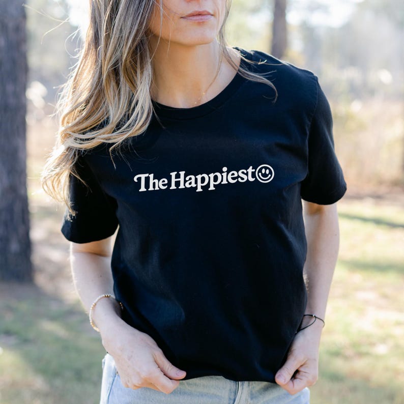 Puede incluir: Una camiseta negra de manga corta con el texto blanco The Happiest seguido de un icono de cara sonriente blanca centrado en el pecho. La prenda tiene un cuello redondo cl&aacute;sico y un dobladillo recto, ofreciendo un dise&ntilde;o sencillo y limpio.