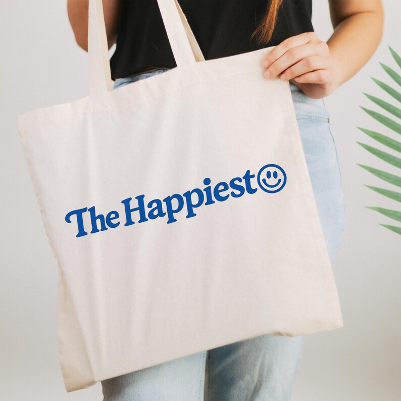 Puede incluir: Un bolso de tela color crema con el texto The Happiest en una fuente serif azul negrita, acompa&ntilde;ado de un icono de cara sonriente azul al final de la frase. El bolso tiene dos asas de tela a juego y una forma rectangular sencilla.