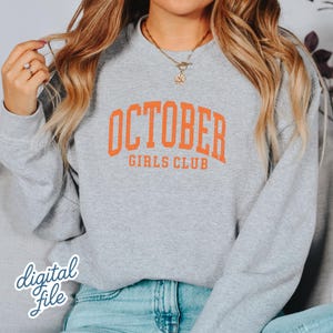 May include: A heather gray crewneck sweatshirt featuring the text OCTOBER GIRLS CLUB printed in a bold orange collegiate font across the chest. The garment has long sleeves and a ribbed neckline, providing a classic fit for casual wear.