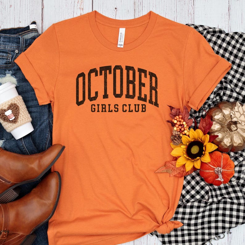 May include: An orange short-sleeved t-shirt with the black text OCTOBER GIRLS CLUB printed on the front. The shirt is displayed alongside blue denim jeans, brown leather boots, a coffee cup in a knit sleeve, a sunflower, and a small pumpkin.