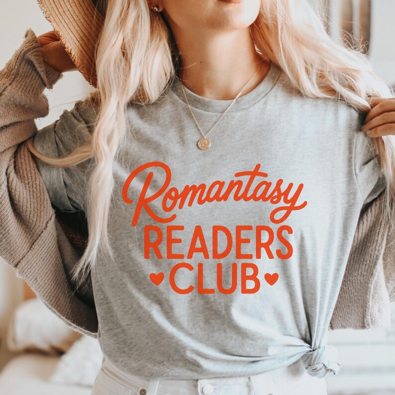 May include: A light gray t-shirt with the words "Romantasy READERS CLUB" in red script and block letters. Two red hearts are below the word "CLUB". The shirt is made of a soft material.