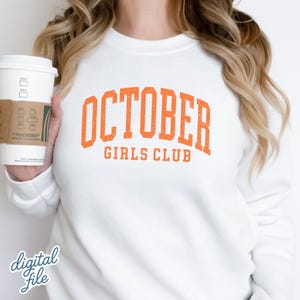 May include: A white crewneck sweatshirt featuring the orange text OCTOBER GIRLS CLUB in a distressed collegiate font across the chest. The garment is shown in a plain white color with long sleeves and a ribbed neckline, presented as a clean design.