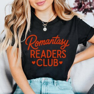 May include: Black t-shirt with the words "Romantasy READERS CLUB" in red script. The text is centered on the chest. Two red hearts are below the word "CLUB". The model is wearing blue jeans and a gold necklace.