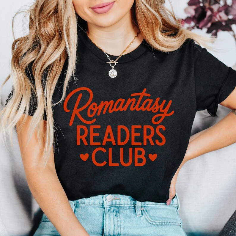 May include: Black t-shirt with the words "Romantasy READERS CLUB" in red script. The text is centered on the chest. Two red hearts are below the word "CLUB". The model is wearing blue jeans and a gold necklace.