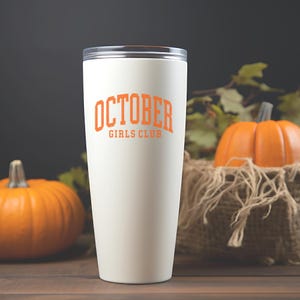 May include: A white stainless steel tumbler featuring the orange text OCTOBER GIRLS CLUB printed on the front. The container has a clear lid and a tapered cylindrical shape, standing upright with a smooth matte finish and a clean design.