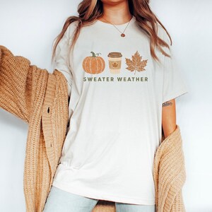Puede incluir: Una camiseta de manga corta color crema con un estampado gr&aacute;fico de una calabaza naranja, un vaso de caf&eacute; beige y una hoja de arce marr&oacute;n. Debajo de los iconos, el texto "SWEATER WEATHER" aparece escrito en letras may&uacute;sculas de color verde.