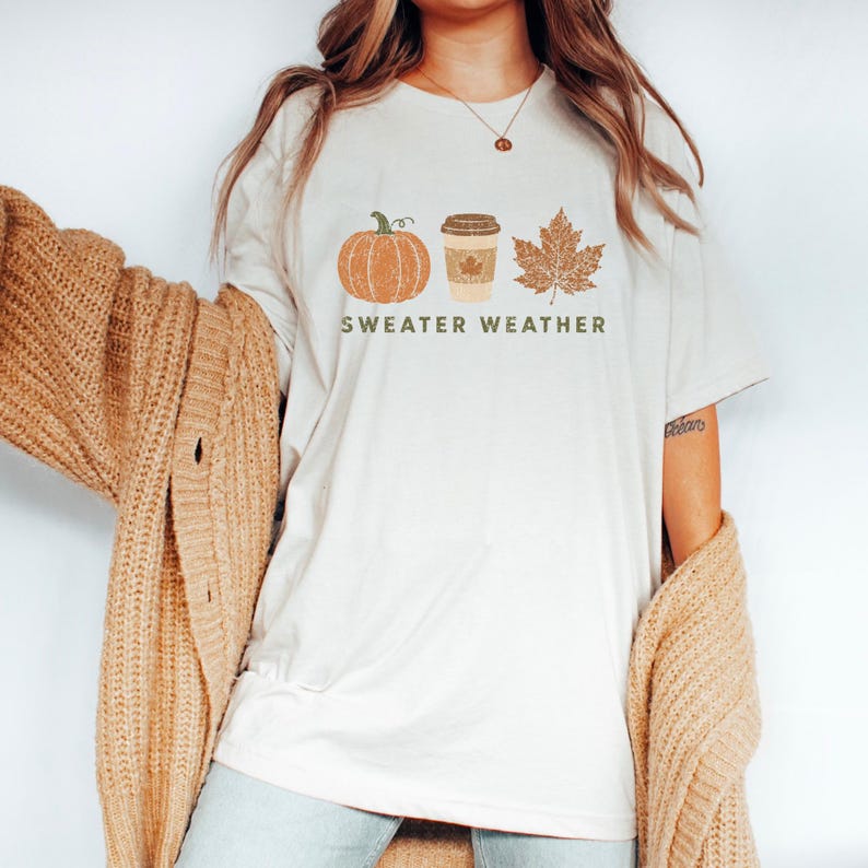 Puede incluir: Una camiseta de manga corta color crema con un estampado gr&aacute;fico de una calabaza naranja, un vaso de caf&eacute; beige y una hoja de arce marr&oacute;n. Debajo de los iconos, el texto "SWEATER WEATHER" aparece escrito en letras may&uacute;sculas de color verde.