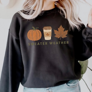 Puede incluir: Una sudadera negra de cuello redondo con un estampado gr&aacute;fico de una calabaza naranja, un vaso de caf&eacute; beige y una hoja de arce marr&oacute;n. Debajo de los iconos, el texto "SWEATER WEATHER" est&aacute; impreso en color verde oliva con textura.