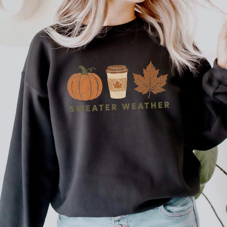 Puede incluir: Una sudadera negra de cuello redondo con un estampado gr&aacute;fico de una calabaza naranja, un vaso de caf&eacute; beige y una hoja de arce marr&oacute;n. Debajo de los iconos, el texto "SWEATER WEATHER" est&aacute; impreso en color verde oliva con textura.