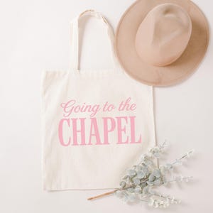 Puede incluir: Una bolsa de tela color crema con el texto rosa Going to the CHAPEL impreso en la parte delantera. La bolsa se acompa&ntilde;a de un sombrero beige de ala ancha y una rama de hojas de eucalipto verdes, creando un conjunto de accesorios coordinado.