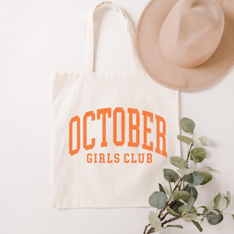 May include: A cream-colored canvas tote bag featuring the text OCTOBER GIRLS CLUB printed in a distressed orange collegiate font. The bag has two shoulder straps and is accompanied by a beige felt hat and a sprig of green eucalyptus leaves.