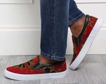 May include: Slip-on sneakers with a vibrant red, yellow, and black patterned fabric upper. The shoes have black leather accents and white soles. The person is wearing blue denim jeans.