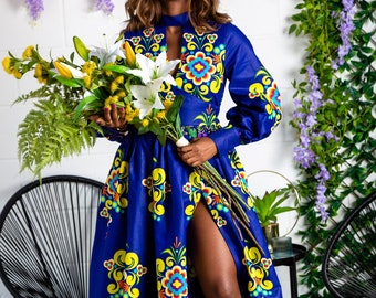 May include: A woman in a long, blue dress with a floral pattern, holding a bouquet of white lilies and yellow flowers. The dress has a high slit and long sleeves. The background includes greenery and a black chair.