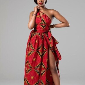 May include: A woman wearing a red and gold patterned maxi dress with a thigh-high slit. The dress has a one-shoulder design and a belt.