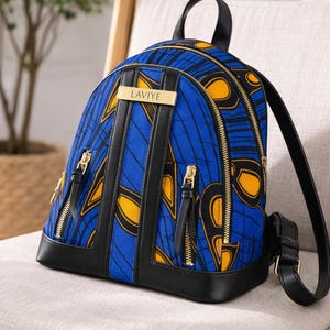 May include: A blue and yellow patterned backpack with black leather accents. The backpack has gold zippers and a small gold nameplate that reads "LAVIYE". The bag is sitting on a cream-colored chair.