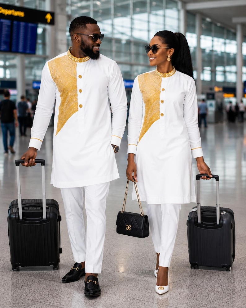 May include: Matching white long-sleeve tunics with gold textured panels on the chest, gold-trimmed collars, and gold buttons. The outfits are paired with white trousers, black loafers, and white heels, accompanied by black rolling suitcases and a black handbag.