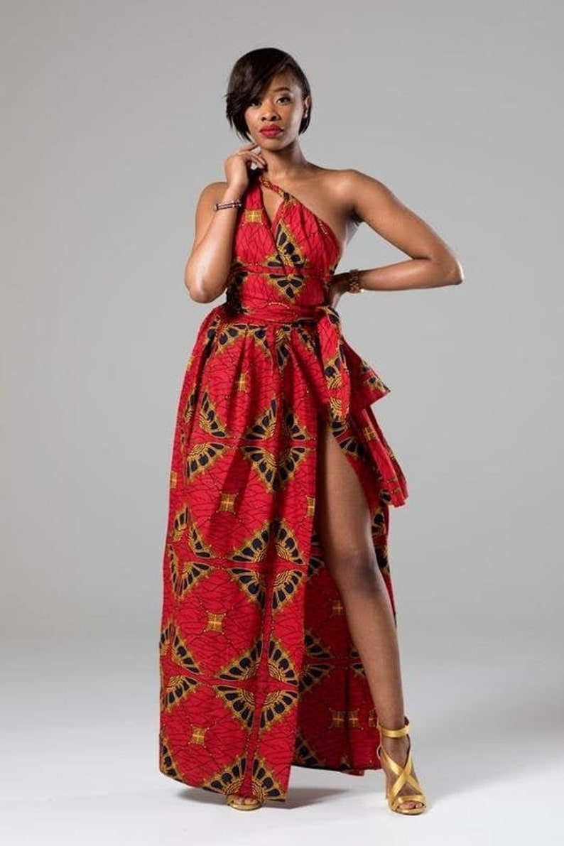May include: A woman wearing a red and gold patterned maxi dress with a thigh-high slit. The dress has a one-shoulder design and a belt.