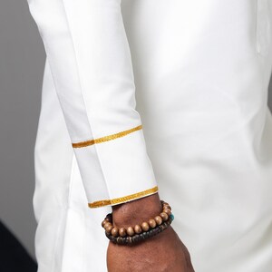 May include: A long-sleeved white shirt featuring gold embroidered trim on the cuffs and a matching gold embroidered detail on the upper chest area. The garment is worn with two stacked beaded bracelets made of dark brown wooden spheres.