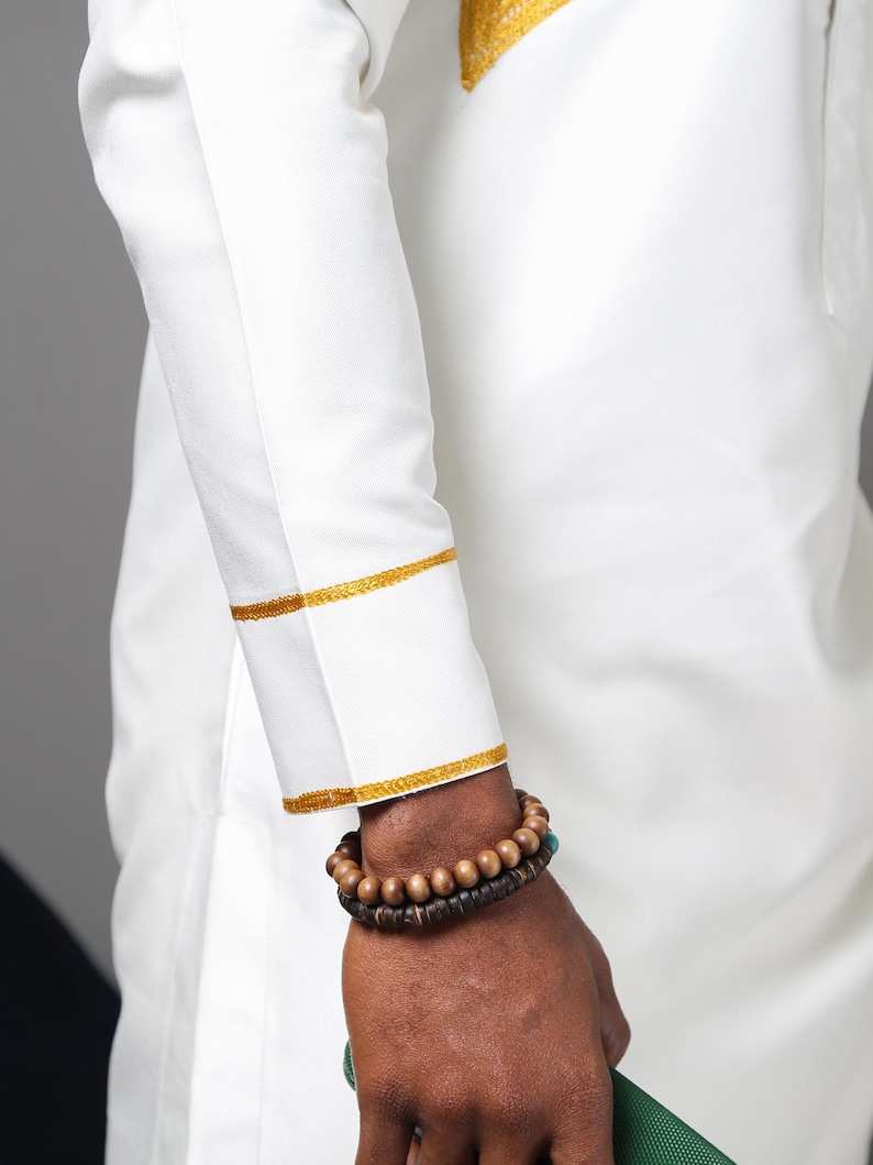 May include: A long-sleeved white shirt featuring gold embroidered trim on the cuffs and a matching gold embroidered detail on the upper chest area. The garment is worn with two stacked beaded bracelets made of dark brown wooden spheres.