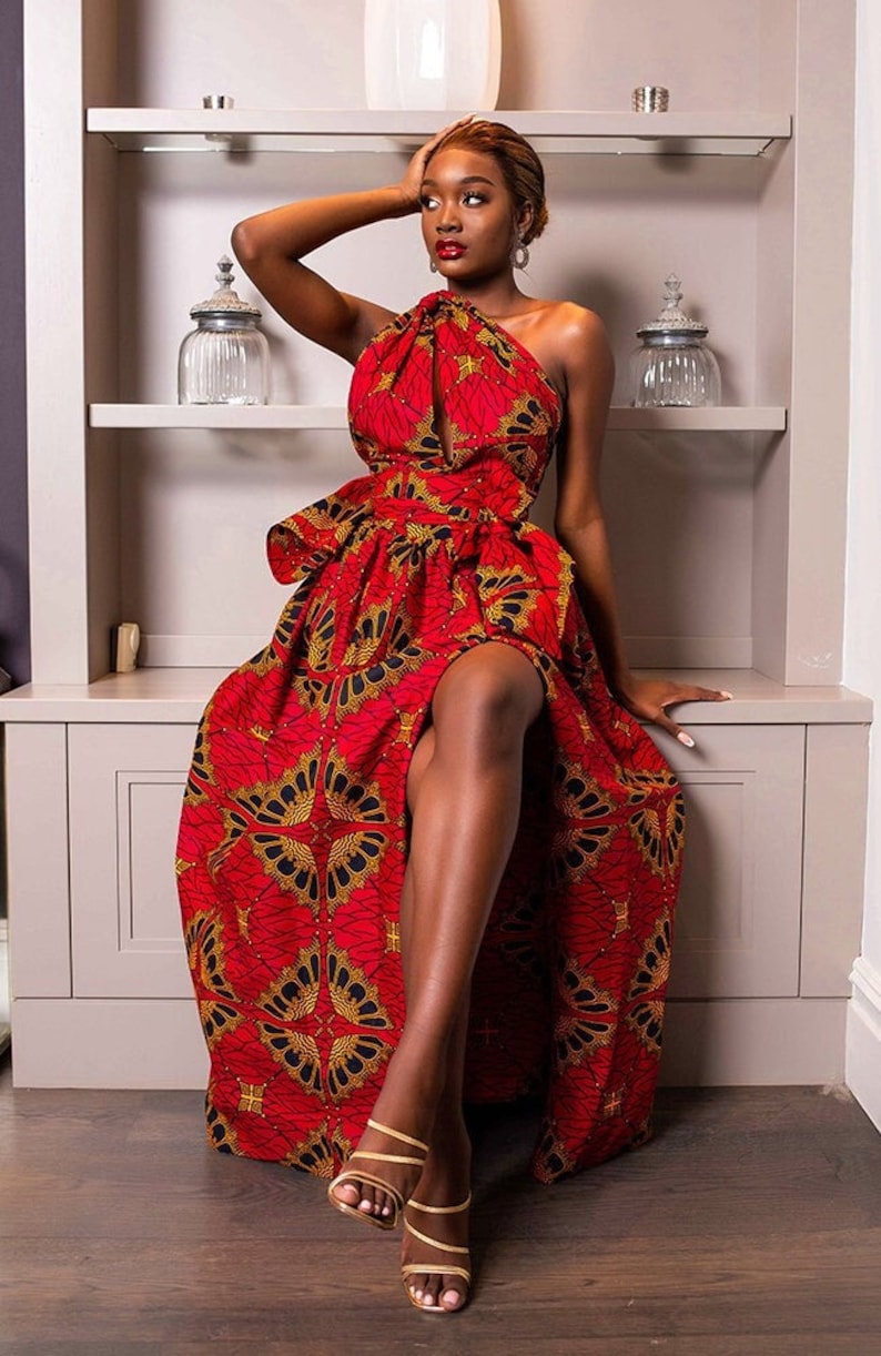 May include: A woman wearing a red and gold patterned maxi dress with a one-shoulder neckline and a high slit. She is sitting on a white cabinet with a shelf behind her.