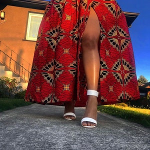 May include: A red maxi skirt with a black and gold geometric pattern and a high slit. The skirt is worn with white heeled sandals.
