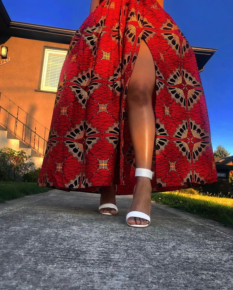 May include: A red maxi skirt with a black and gold geometric pattern and a high slit. The skirt is worn with white heeled sandals.