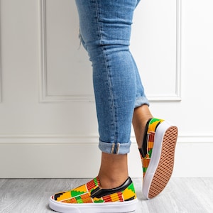May include: Slip-on sneakers with a vibrant African print in red, yellow, green, and black. The shoes have a white sole and a black leather trim. The sneakers are paired with blue jeans.