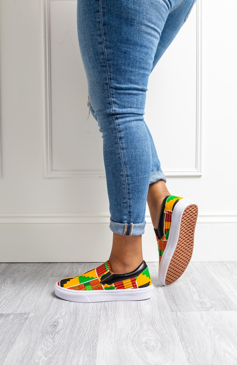 May include: Slip-on sneakers with a vibrant African print in red, yellow, green, and black. The shoes have a white sole and a black leather trim. The sneakers are paired with blue jeans.