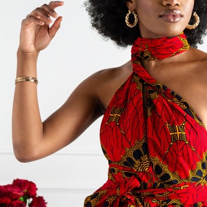May include: A woman wearing a red and gold patterned halter dress with a black belt. The dress has a wrap-style design and a high neckline. The woman has an afro hairstyle and is wearing gold hoop earrings.