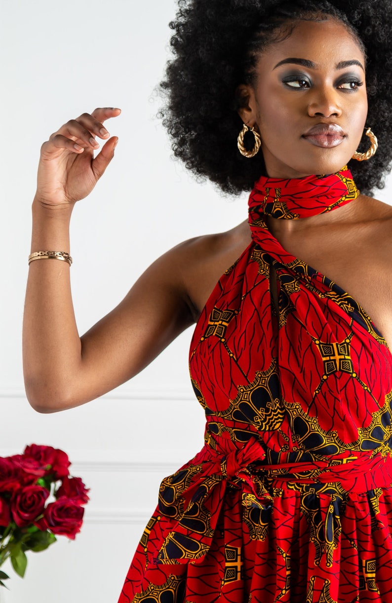 May include: A woman wearing a red and gold patterned halter dress with a black belt. The dress has a wrap-style design and a high neckline. The woman has an afro hairstyle and is wearing gold hoop earrings.