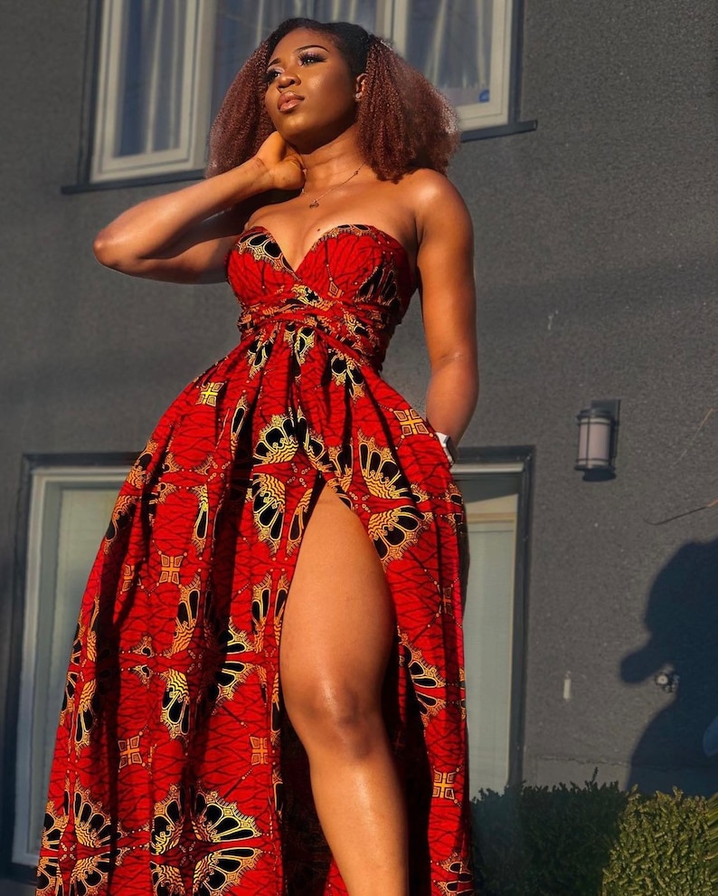May include: A woman wearing a red and gold strapless maxi dress with a thigh-high slit. The dress has a bold African print pattern.