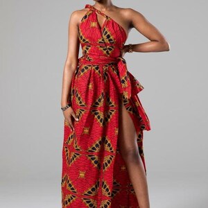 May include: A red maxi dress with a gold and black geometric print. The dress has a one-shoulder neckline and a thigh-high slit on the left side. The dress is cinched at the waist with a matching fabric belt.