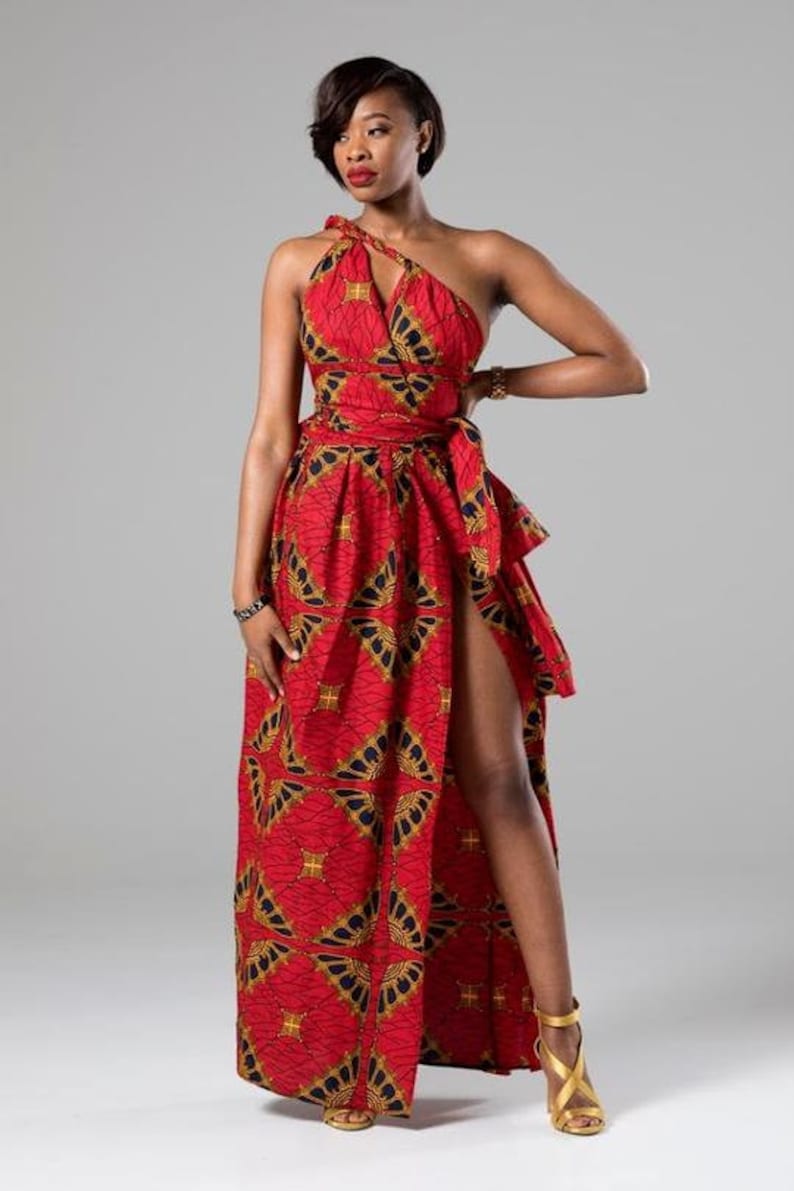 May include: A red maxi dress with a gold and black geometric print. The dress has a one-shoulder neckline and a thigh-high slit on the left side. The dress is cinched at the waist with a matching fabric belt.