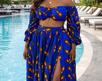 May include: A two-piece set consisting of an off-the-shoulder crop top with long puff sleeves and a matching high-waisted maxi skirt featuring a high side slit. The fabric is a vibrant royal blue with a repeating orange and black teardrop pattern.