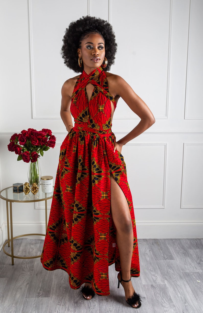 May include: A red maxi dress with a black and gold print. The dress has a halter neckline and a thigh-high slit. The model is wearing the dress with black heels.