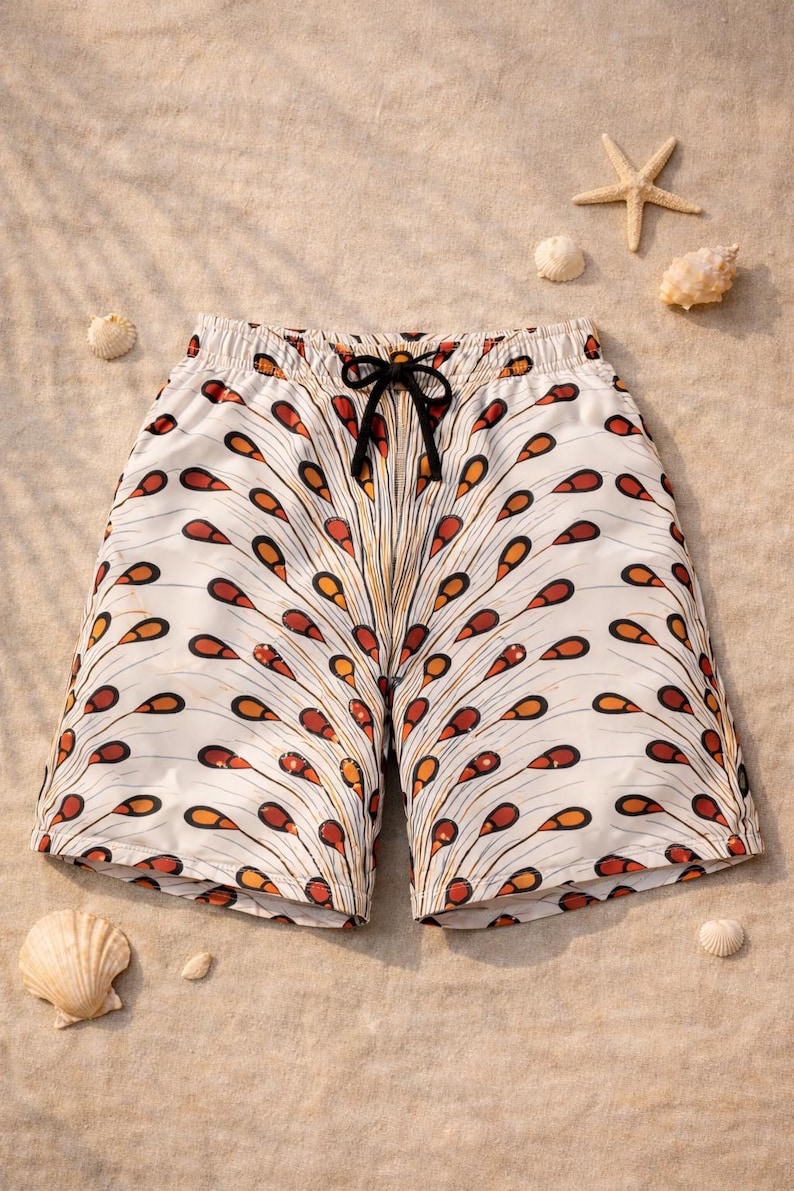 May include: White swim shorts featuring a repeating pattern of orange and brown teardrop shapes. The shorts have a black drawstring and are displayed on a sandy surface with seashells and a starfish.