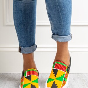 May include: Slip-on sneakers with a vibrant African print in yellow, red, green, and black. The shoes have a white sole and black trim. The person is wearing blue jeans with distressed details and rolled-up cuffs.