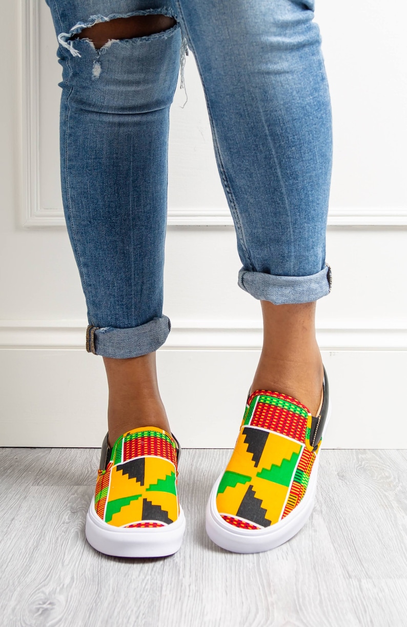 May include: Slip-on sneakers with a vibrant African print in yellow, red, green, and black. The shoes have a white sole and black trim. The person is wearing blue jeans with distressed details and rolled-up cuffs.