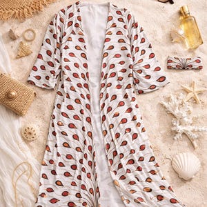 May include: A white kimono with a red and orange teardrop design, displayed on sand with a straw hat, sunglasses, and accessories. The lightweight, flowing garment is ideal for beachwear or a summer cover-up. The kimono's length is approximately 130 cm.
